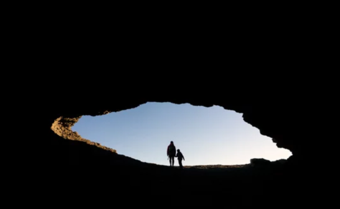 Madre e hijo con chaquetas rojas silueteados en la cueva de La ojerada en la costa del norte de España al atardecer, Ajo, Cantabria.