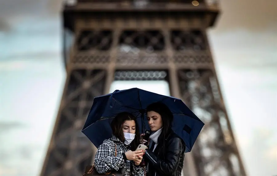 Dos mujeres cobijadas bajo un paraguas observan un tel&eacute;fono m&oacute;vil frente a la Torre Eiffel, este lunes en Par&iacute;s (Francia).EFE/ Ian Langsdon