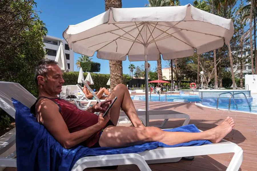 Tom E., trabajador en tecnolog&iacute;as de la informaci&oacute;n trabaja con su tablet mientras toma el sol en la piscina del hotel "Playa del Sol", en la localidad tur&iacute;stica de Playa del Ingl&eacute;s (Gran Canaria), donde se aloja, establecimiento que tiene el cartel de completo gracias a su reconversi&oacute;n en alojamiento para trabajadores remotos. Algunos hoteles de Gran Canaria se han reinventado y mientras muchos echan el cierre a causa del cero tur&iacute;stico de la pandemia, otros como "Playa del Sol" han colgado por sorpresa el cartel de completo gracias a una nueva receta que mezcla el ocio y el trabajo con un nuevo concepto hotelero. EFE/&Aacute;ngel Medina G.