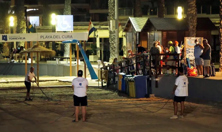 Varias personas (d) hacen cola ante el acceso a la playa de madrugada para coger sitio mientras operarios municipales parcelan la arena para evitar la concentraci&oacute;n de veraneantes y evitar la propagaci&oacute;n del coronavirus, este s&aacute;bado de madrugada en Torrevieja. EFE / Manuel Lorenzo