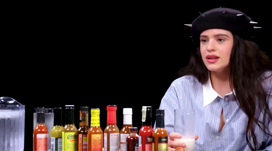La cantante Rosalia, con un vaso de leche en la mano, y los botes de las salsas picantes. / Hot Ones