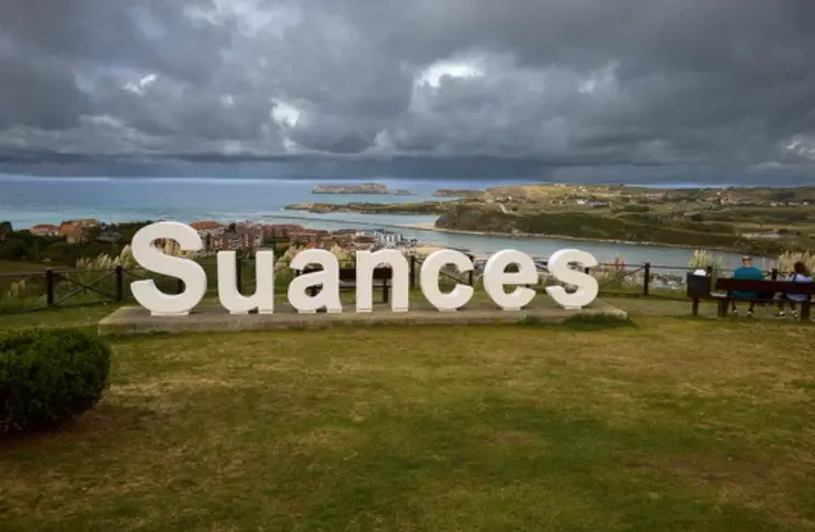 Dos personas junto al cartel de 'Suances' bajo un manto de nubes.