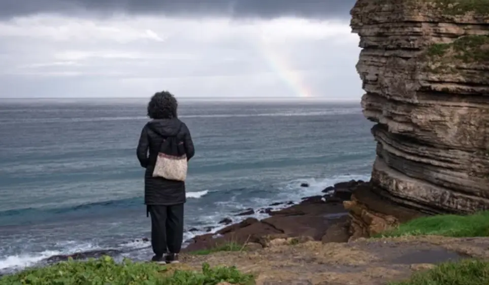 Una mujer contempla el mar Cant&aacute;brico desde un acantilado, mientras un arco&iacute;ris asoma en el horizonte tras el paso de la tormenta.