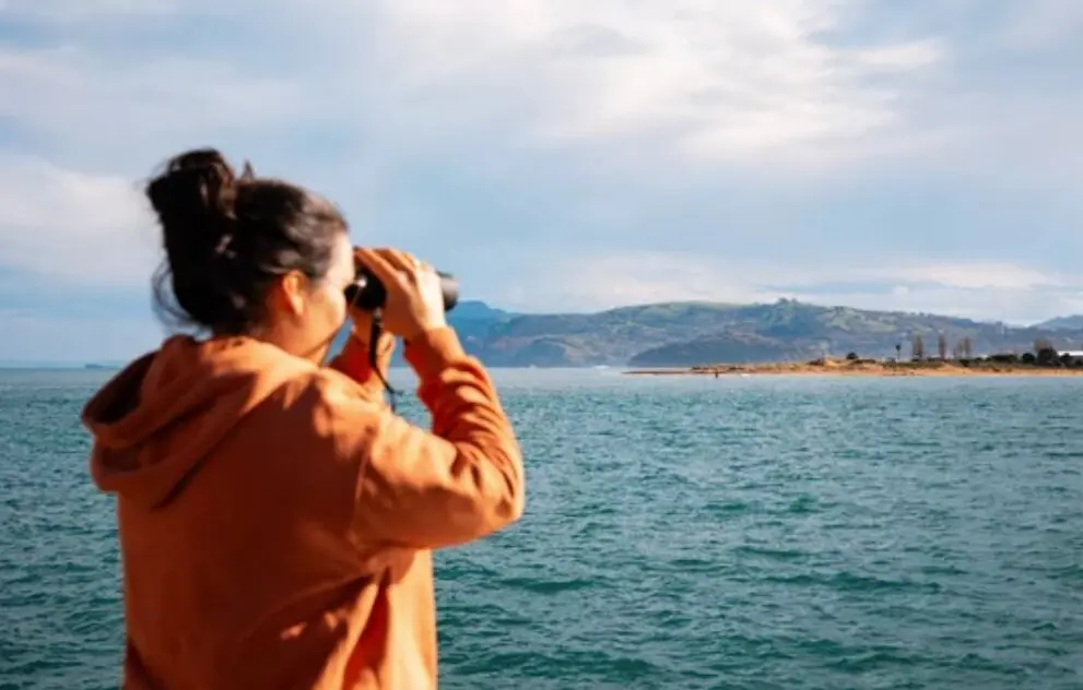 Una mujer observa el horizonte costero de Cantabria con prism&aacute;ticos desde una embarcaci&oacute;n, aprovechando un claro entre nubes tras las lluvias matinales.