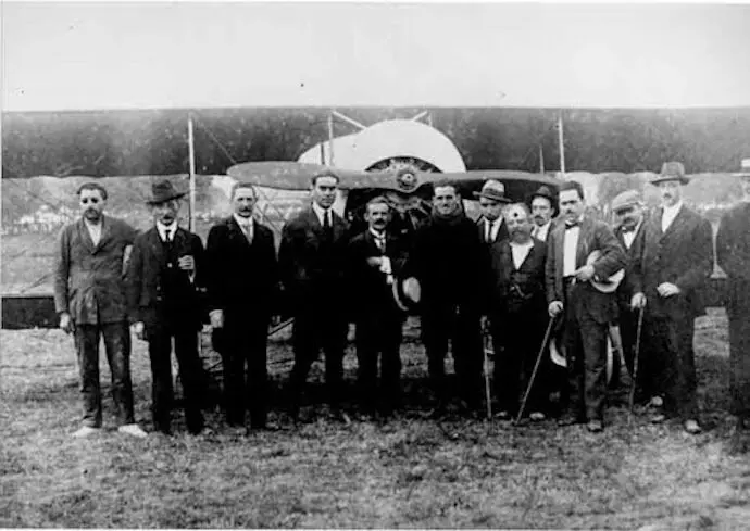 Joaqu&iacute;n Cay&oacute;n (centro con sombrero) rodeado de autoridades y vecinos tras su vuelo hist&oacute;rico sobre Torrelavega en 1921, con su rudimentario avi&oacute;n construido con materiales reciclados al fondo. La imagen refleja la admiraci&oacute;n popular por este pionero torrelaveguense de la aviaci&oacute;n.