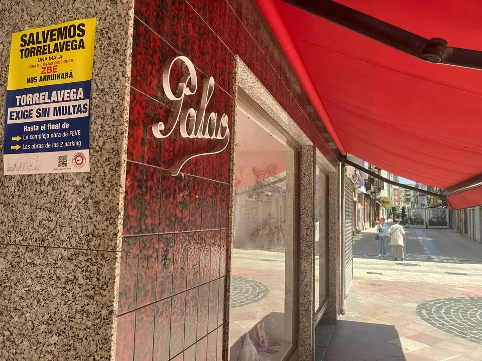 Cartel de protesta contra las multas de la ZBE en un comercio cerrado del centro de Torrelavega, mientras dos vecinas caminan por una calle semivac&iacute;a marcada por las obras y la crisis comercial.
