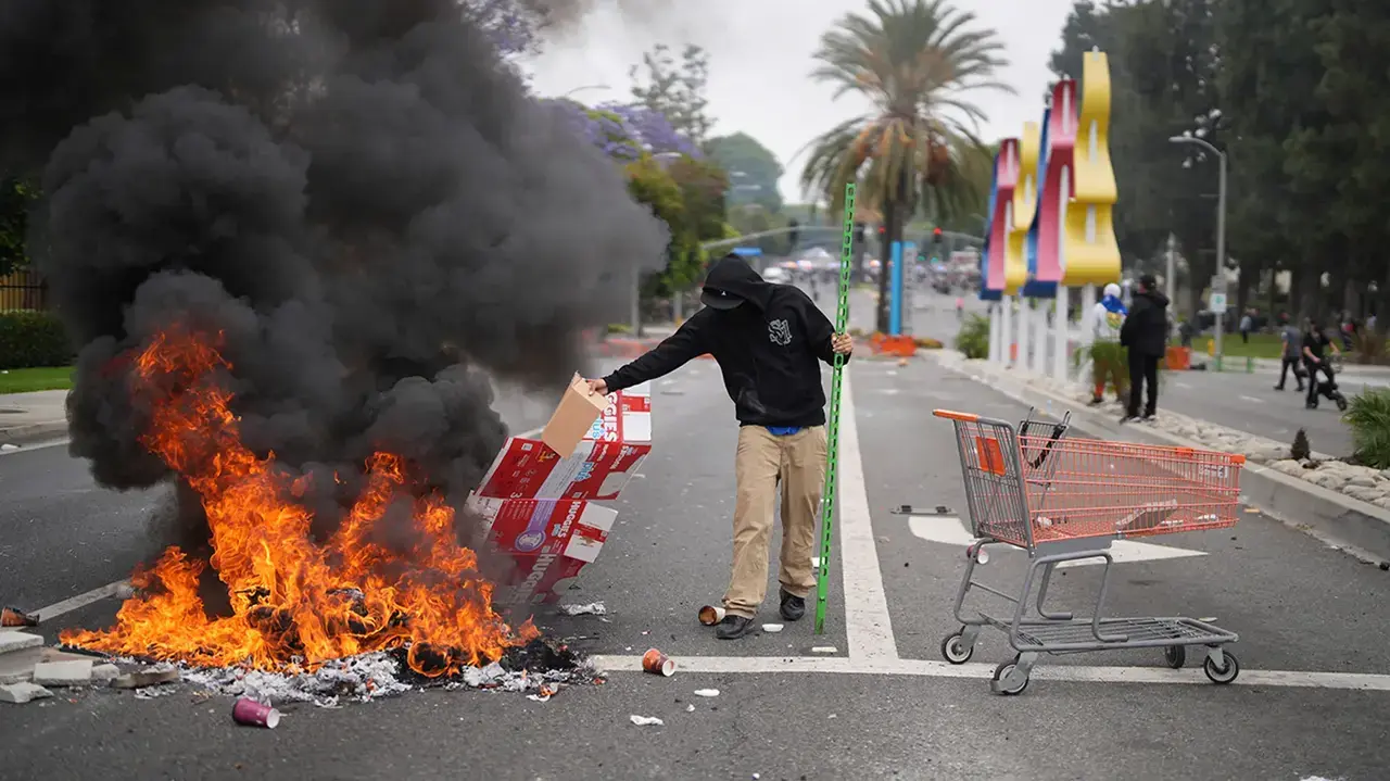 Un manifestante coloca escombros en una hoguera mientras personal de la Patrulla Fronteriza con equipo antidisturbios y m&aacute;scaras de gas monta guardia afuera de un parque industrial en Paramount, California, el s&aacute;bado 7 de junio de 2025.  (Foto AP/Eric Thayer)