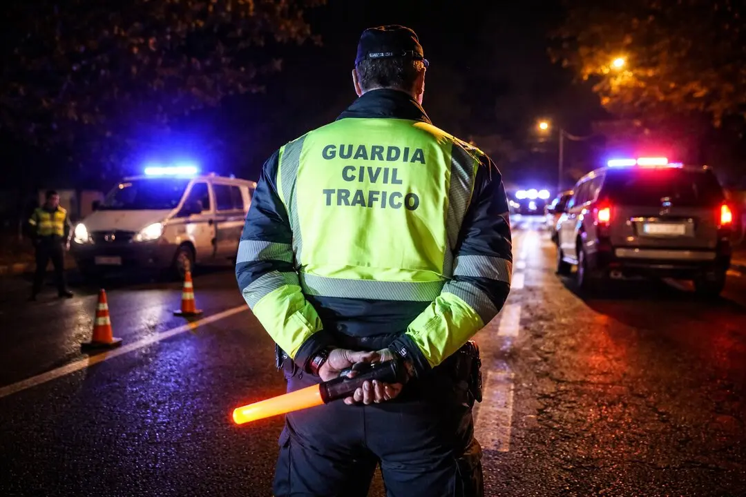 Un agente de la Guardia Civil de Tr&aacute;fico supervisa un control nocturno en carretera, con veh&iacute;culos oficiales iluminando la escena.