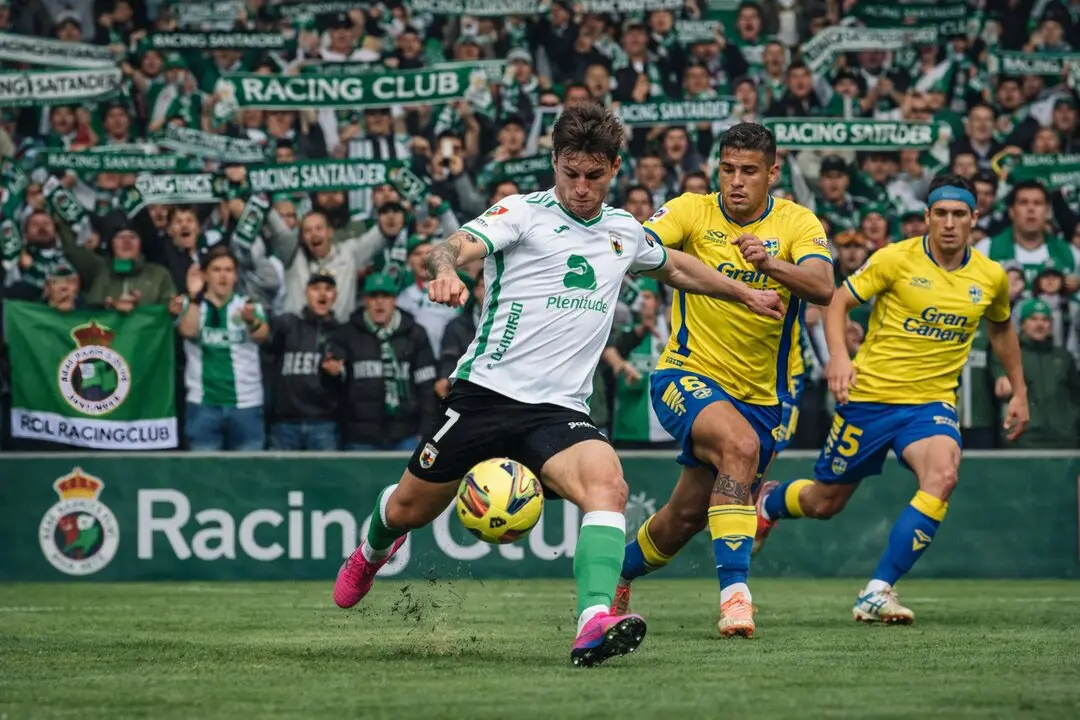 Duelo intenso entre el Racing de Santander y la UD Las Palmas: el jugador n&uacute;mero 7 del Racing controla el bal&oacute;n ante la presi&oacute;n rival, mientras la afici&oacute;n c&aacute;ntabra anima con fuerza desde la grada del estadio.