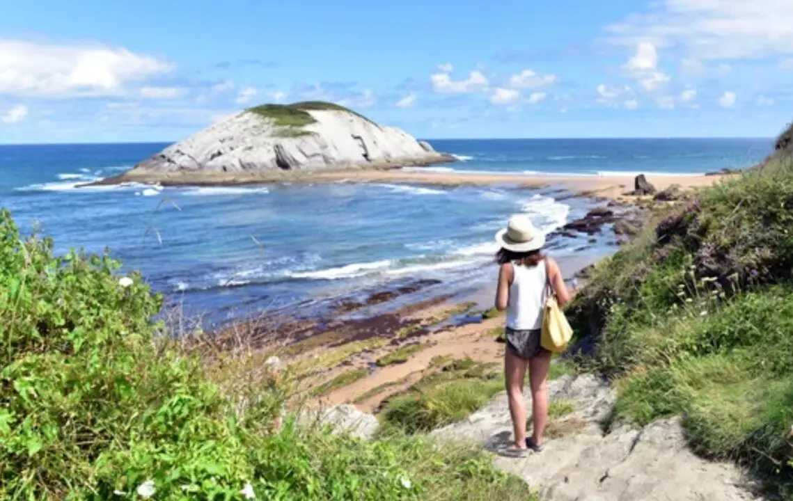 Una visitante contempla la costa c&aacute;ntabra en una jornada soleada, con el mar en calma y cielo despejado frente a un islote del litoral.