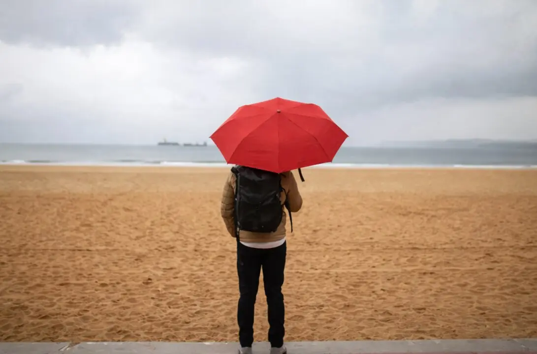 Una persona con paraguas rojo en la playa de El Sardinero, en Santander, en una jornada marcada por el cielo gris y la lluvia.