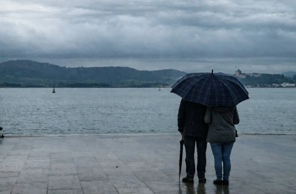 Una pareja contempla la bah&iacute;a de Santander bajo un paraguas mientras la lluvia y las nubes bajas envuelven el paisaje en una tarde gris.