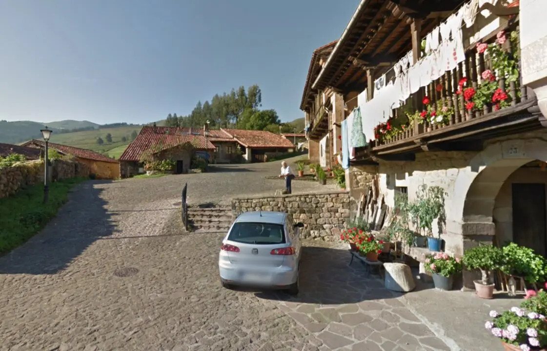 Un vecino descansa en la plaza empedrada de Carmona (Cantabria), rodeado de casas monta&ntilde;esas con balconadas de madera y flores, en una escena cotidiana que refleja la vida tranquila del valle de Cabu&eacute;rniga.