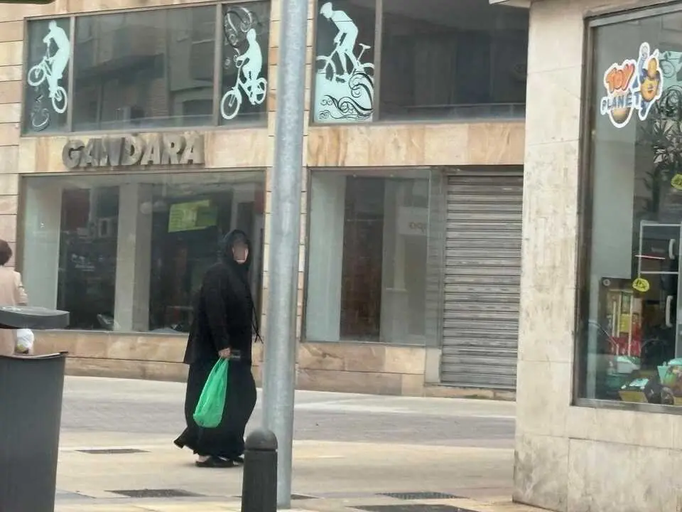 Una mujer camina por una calle comercial, portando una bolsa verde, frente a un establecimiento en el centro urbano.