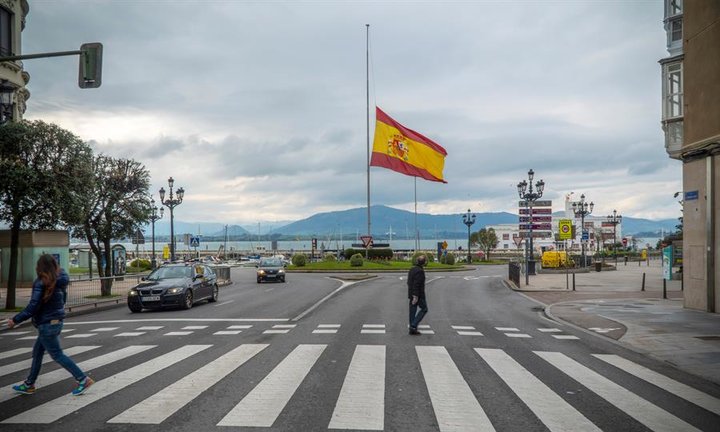 Varios peatones cruzan una calle delante de una bandera que ondea a media asta en memoria de las víctimas de coronavirus, este martes en Santander. EFE/Román G. Aguilera