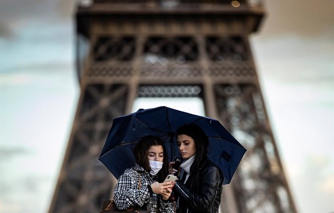 Dos mujeres cobijadas bajo un paraguas observan un teléfono móvil frente a la Torre Eiffel, este lunes en París (Francia).EFE/ Ian Langsdon