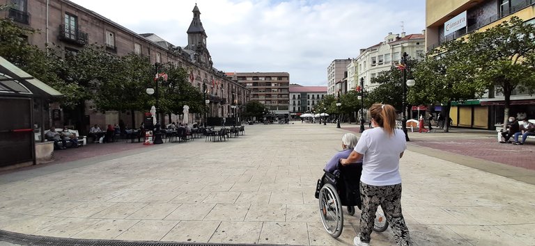 Una persona camina con una persona con movilidad reducida por la plaza del Ayuntamiento de Torrelavega. / S. Díaz