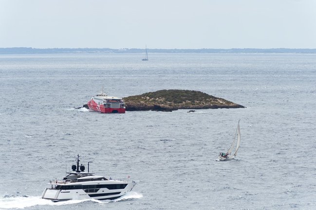 Un barco y un velero pasan cerca del ferry 'San Gwann' de la naviera FRS encallado en el islote norte de Es Malvins, entre Ibiza y Formentera. 
