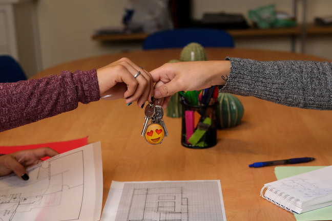 Dos chicas se dan unas llaves de una vivienda. Encima de la mesa hay varios planos sobre viviendas y material de escritura. / Jesús Hellín