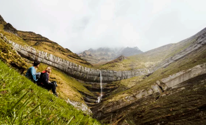 Pareja sentada en la ladera verde de una colina contemplando a una mujer casada que brota de las montañas. Lugar para relajarse y hacer excursiones, situado en Vega de Pas, Cantabria.