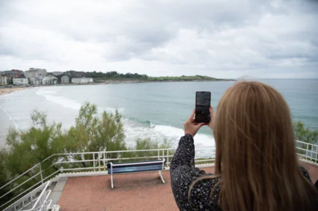 Mujer rubia joven de espaldas sacando una foto al paisaje con su teléfono móvil en la playa de El Sardinero en Santander durante un día nuboso