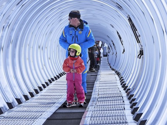 En la estación de esquí de Alto Campoo, un niño acompañado por su monitor asciende por la cinta transportadora, un momento de aprendizaje y aventura en la nieve que encapsula el espíritu del deporte invernal y la transmisión de la pasión por el esquí a las nuevas generaciones.