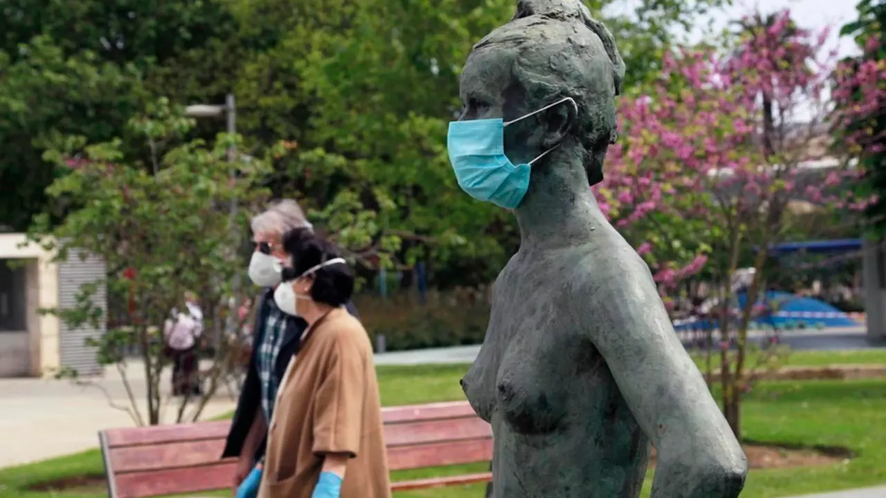 Varias personas con mascarilla caminan junto a una estatua en los jardines de Pereda cubierta tambi&eacute;n por mascarilla. / J. Ram&oacute;n