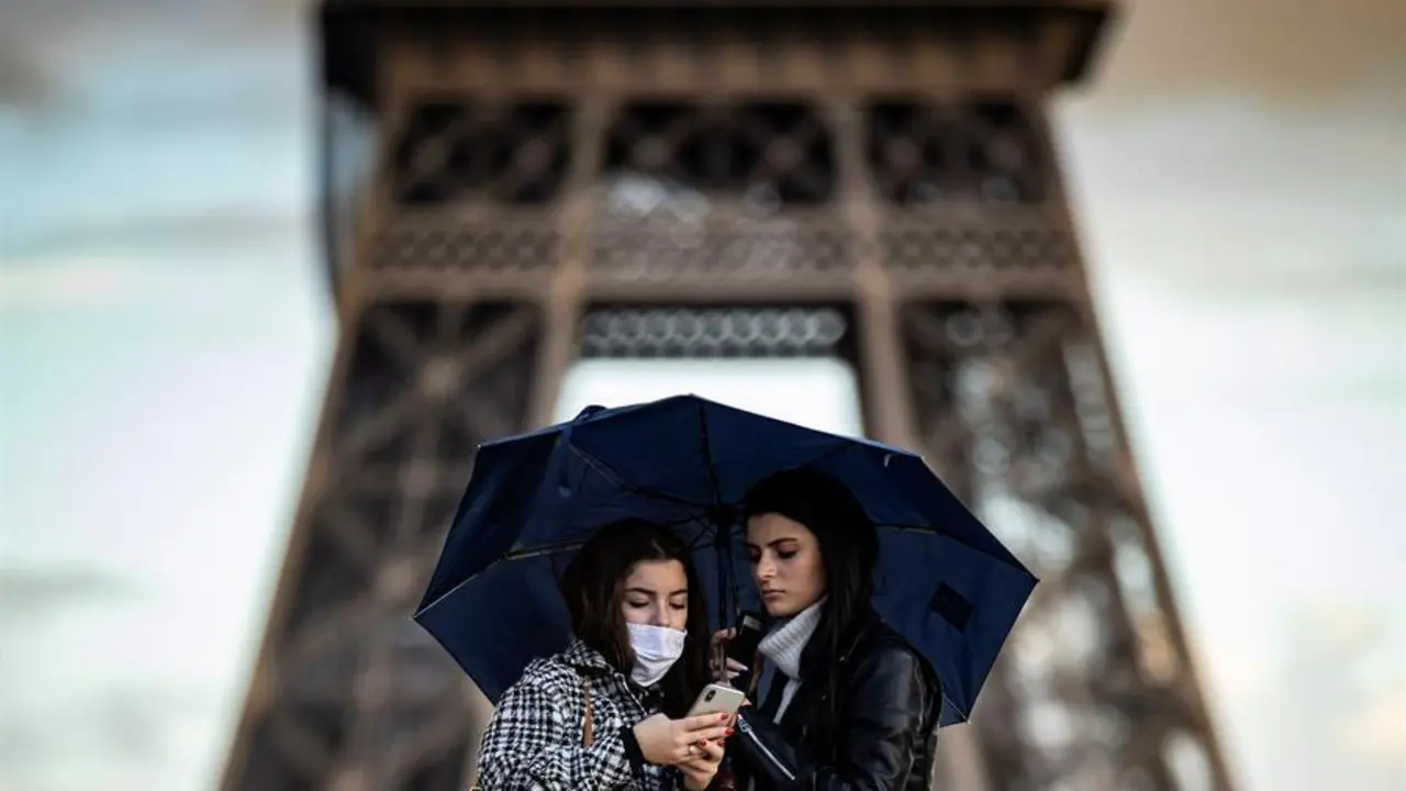 Dos mujeres cobijadas bajo un paraguas observan un tel&eacute;fono m&oacute;vil frente a la Torre Eiffel, este lunes en Par&iacute;s (Francia).EFE/ Ian Langsdon