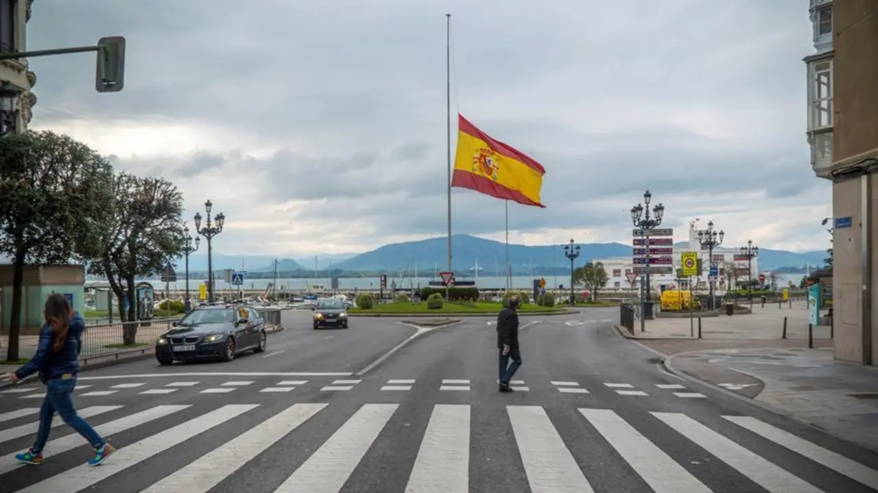 Varios peatones cruzan una calle delante de una bandera que ondea a media asta en memoria de las v&iacute;ctimas de coronavirus, este martes en Santander. /  / luto HARDY / ARCHIVO