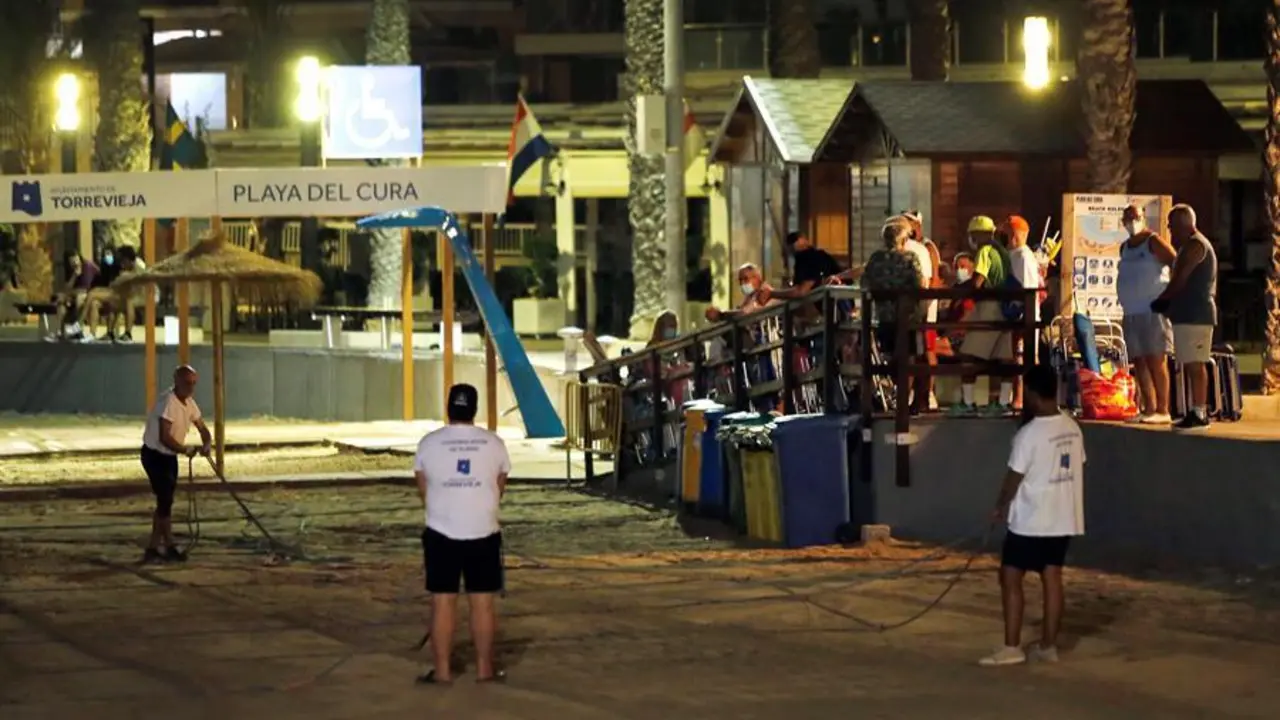 Varias personas (d) hacen cola ante el acceso a la playa de madrugada para coger sitio mientras operarios municipales parcelan la arena para evitar la concentraci&oacute;n de veraneantes y evitar la propagaci&oacute;n del coronavirus, este s&aacute;bado de madrugada en Torrevieja. EFE / Manuel Lorenzo