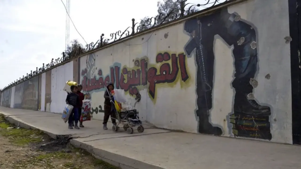 Tres ni&ntilde;os caminan junto a un muro con esl&oacute;ganes del grupo yihadista Estado Isl&aacute;mico (EI)