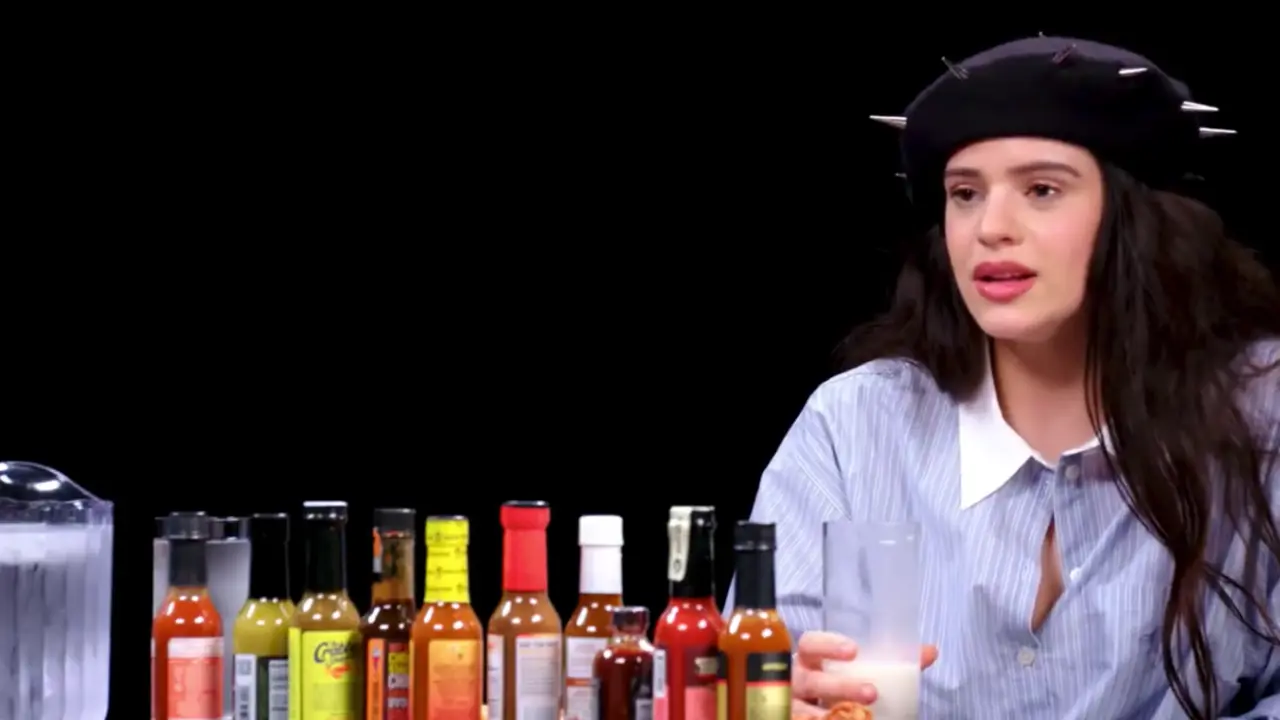 La cantante Rosalia, con un vaso de leche en la mano, y los botes de las salsas picantes. / Hot Ones