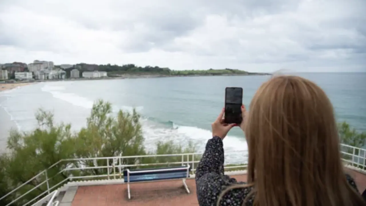 Mujer rubia joven de espaldas sacando una foto al paisaje con su tel&eacute;fono m&oacute;vil en la playa de El Sardinero en Santander durante un d&iacute;a nuboso