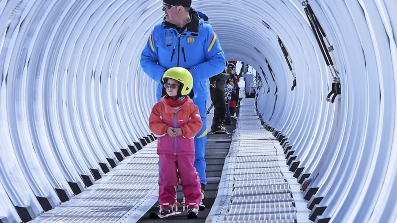 En la estaci&oacute;n de esqu&iacute; de Alto Campoo, un ni&ntilde;o acompa&ntilde;ado por su monitor asciende por la cinta transportadora, un momento de aprendizaje y aventura en la nieve que encapsula el esp&iacute;ritu del deporte invernal y la transmisi&oacute;n de la pasi&oacute;n por el esqu&iacute; a las nuevas generaciones.