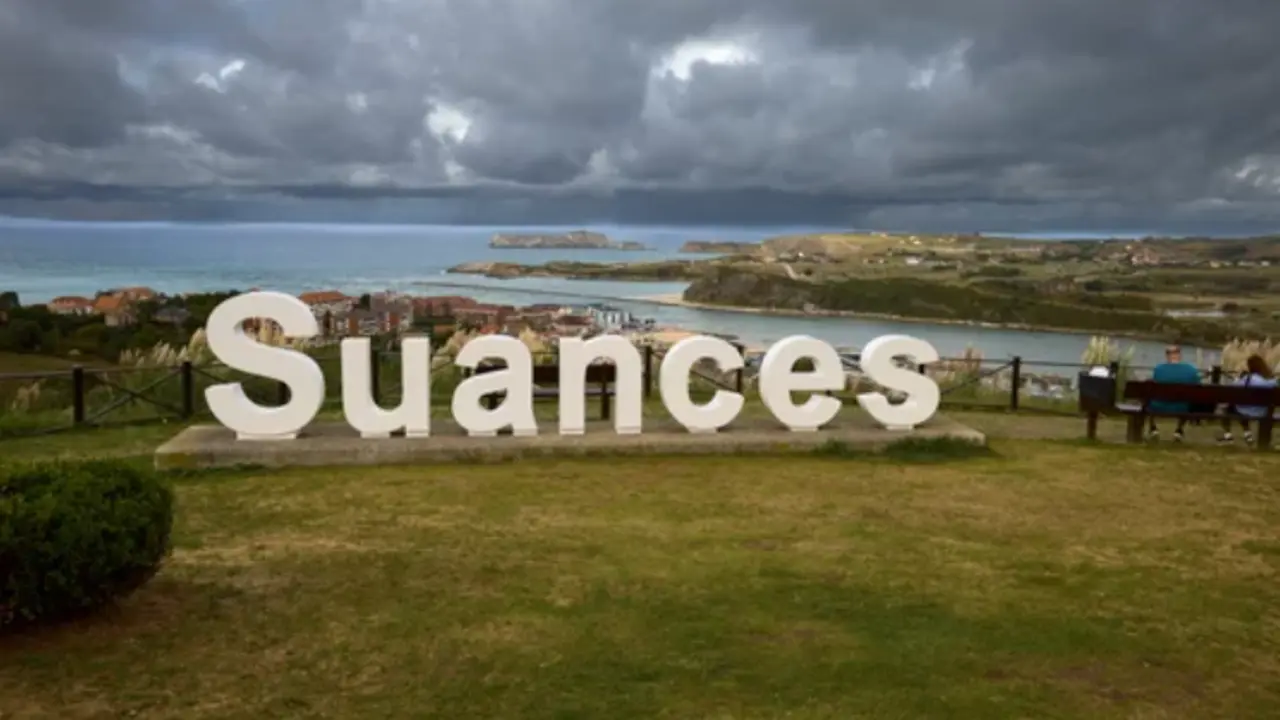 Dos personas junto al cartel de 'Suances' bajo un manto de nubes.
