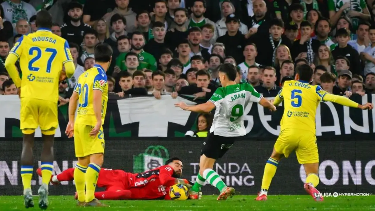 Juan Carlos Arana, delantero del Racing, se lanza con todo en busca del gol mientras el portero del C&aacute;diz y la defensa rival intentan frenar su avance. La afici&oacute;n verdiblanca, de fondo, empuja a su equipo en una noche vibrante de LaLiga Hypermotion.