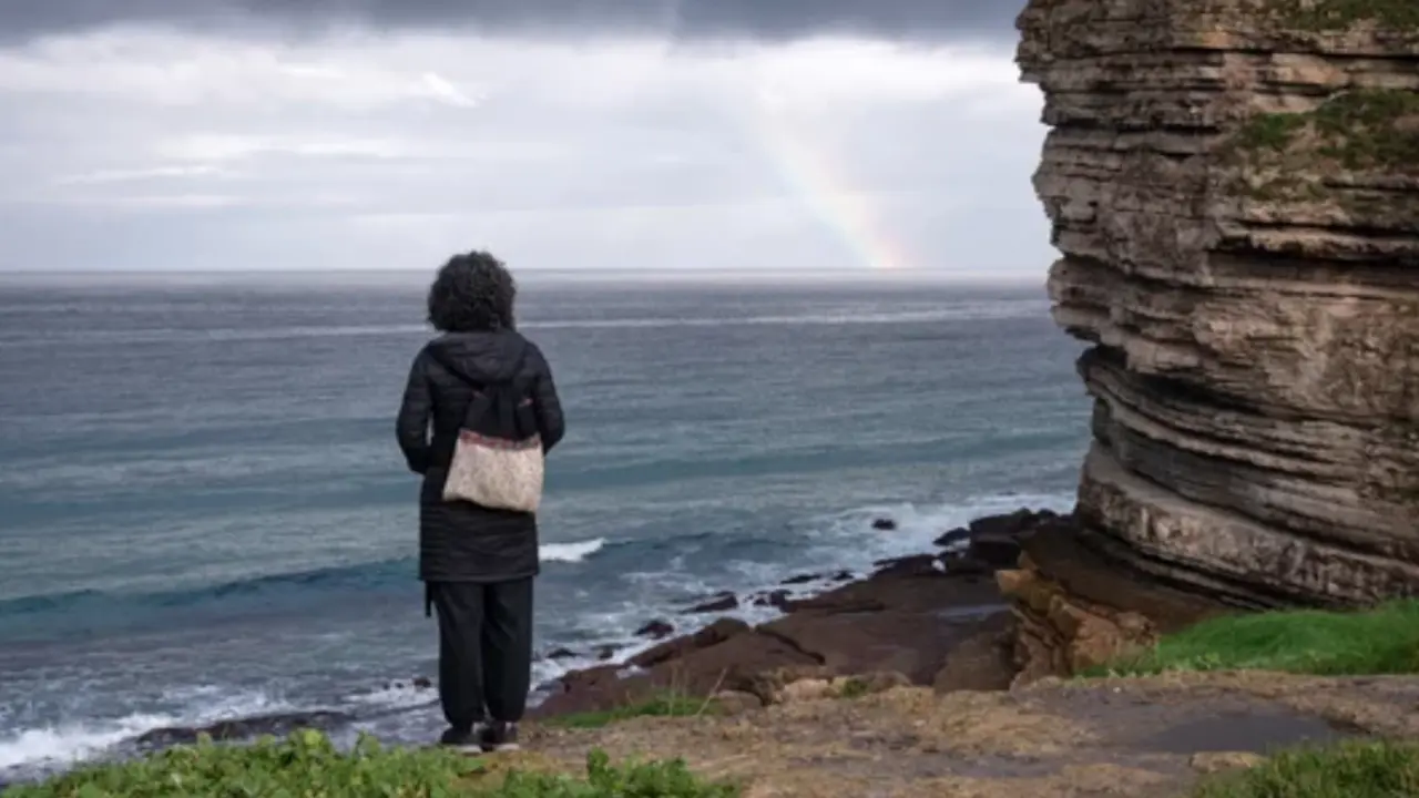 Una mujer contempla el mar Cant&aacute;brico desde un acantilado, mientras un arco&iacute;ris asoma en el horizonte tras el paso de la tormenta.