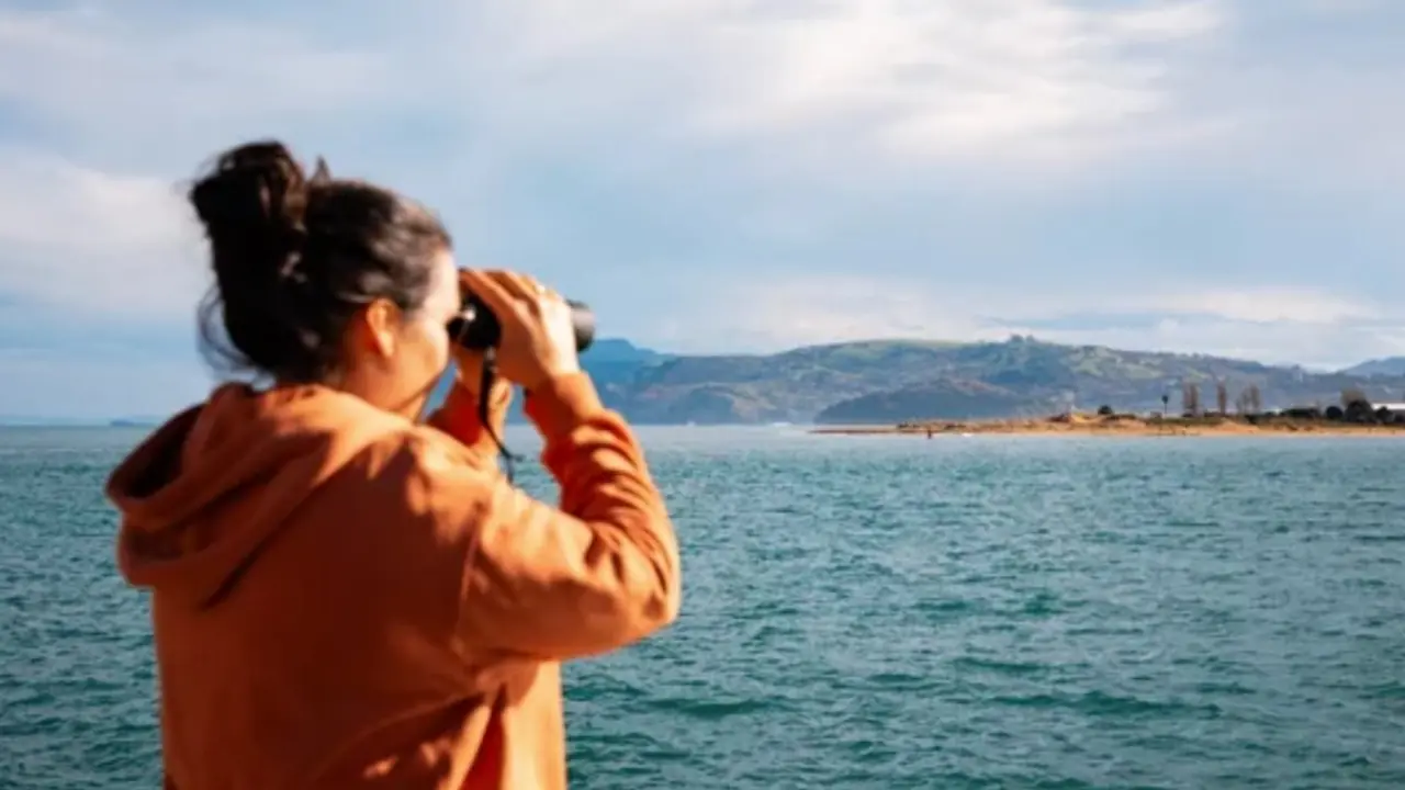 Una mujer observa el horizonte costero de Cantabria con prism&aacute;ticos desde una embarcaci&oacute;n, aprovechando un claro entre nubes tras las lluvias matinales.