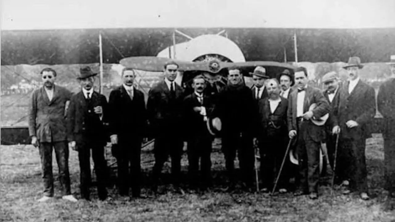 Joaqu&iacute;n Cay&oacute;n (centro con sombrero) rodeado de autoridades y vecinos tras su vuelo hist&oacute;rico sobre Torrelavega en 1921, con su rudimentario avi&oacute;n construido con materiales reciclados al fondo. La imagen refleja la admiraci&oacute;n popular por este pionero torrelaveguense de la aviaci&oacute;n.
