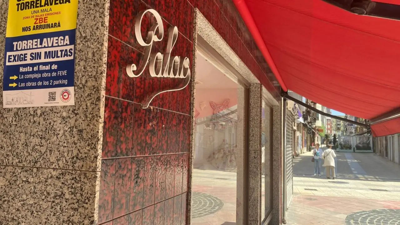 Cartel de protesta contra las multas de la ZBE en un comercio cerrado del centro de Torrelavega, mientras dos vecinas caminan por una calle semivac&iacute;a marcada por las obras y la crisis comercial.