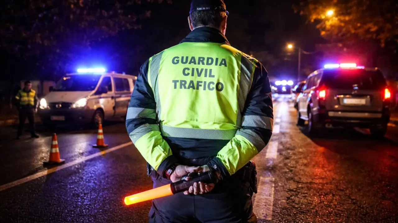 Un agente de la Guardia Civil de Tr&aacute;fico supervisa un control nocturno en carretera, con veh&iacute;culos oficiales iluminando la escena.