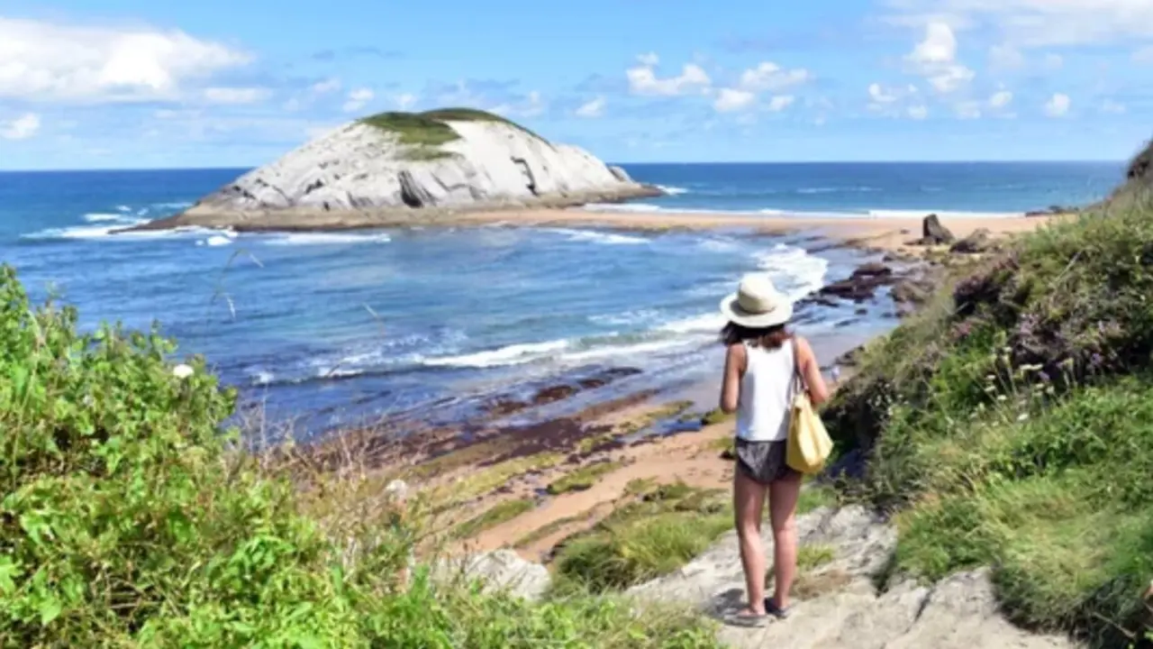 Una visitante contempla la costa c&aacute;ntabra en una jornada soleada, con el mar en calma y cielo despejado frente a un islote del litoral.