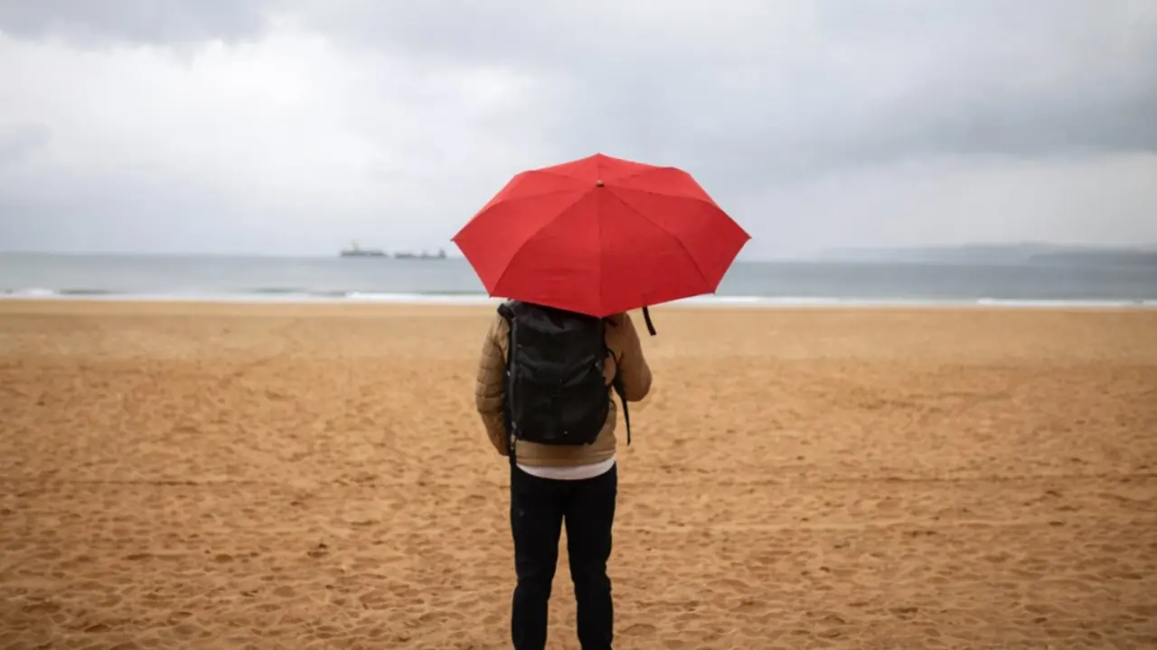 Una persona con paraguas rojo en la playa de El Sardinero, en Santander, en una jornada marcada por el cielo gris y la lluvia.