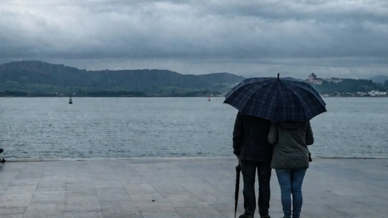 Una pareja contempla la bah&iacute;a de Santander bajo un paraguas mientras la lluvia y las nubes bajas envuelven el paisaje en una tarde gris.