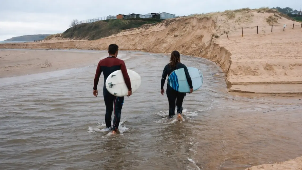 Dos surfistas avanzan por el agua rumbo al mar en una ma&ntilde;ana gris, buscando las primeras olas del d&iacute;a.