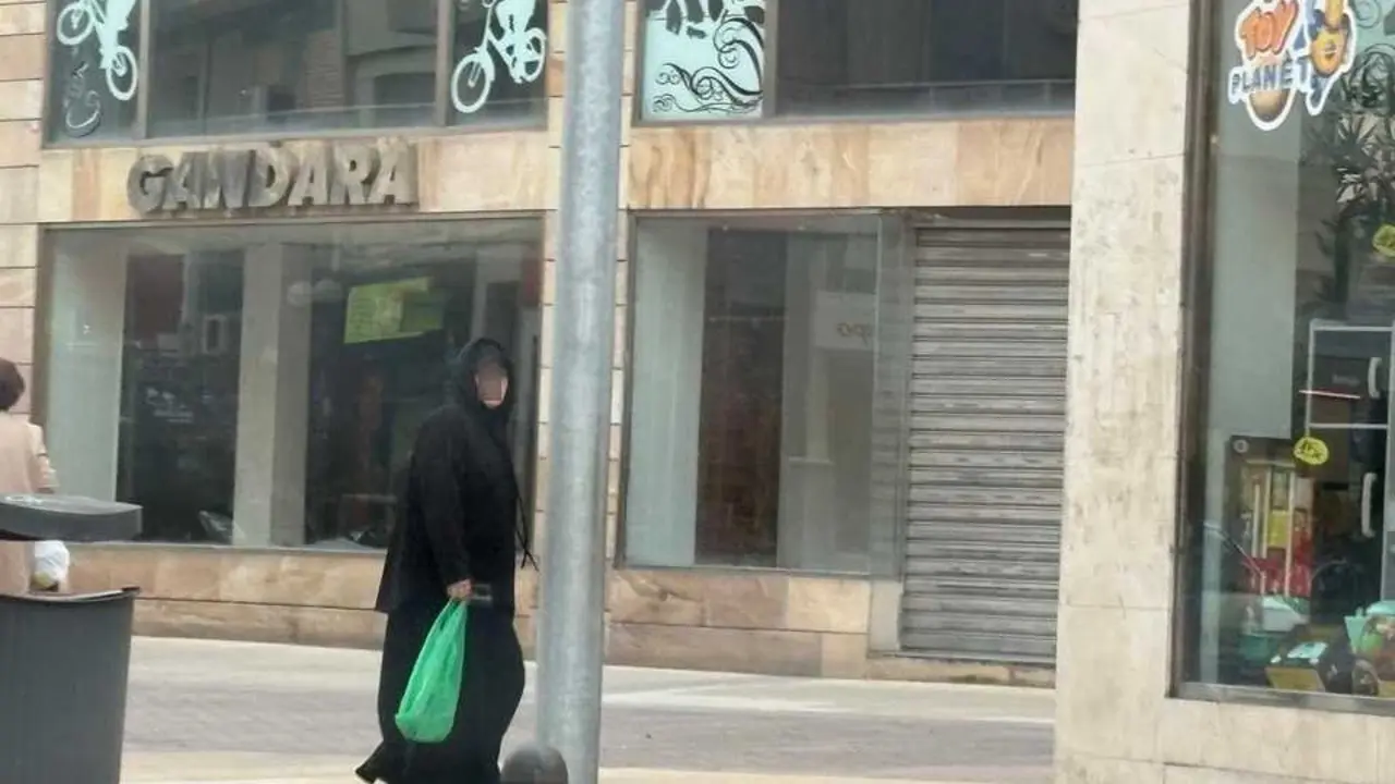 Una mujer camina por una calle comercial, portando una bolsa verde, frente a un establecimiento en el centro urbano.
